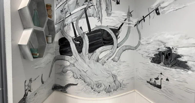 Black and white mural of a giant octopus pulling a pirate ship under the waves, with a skull-shaped island and small boats, painted across the walls of a room